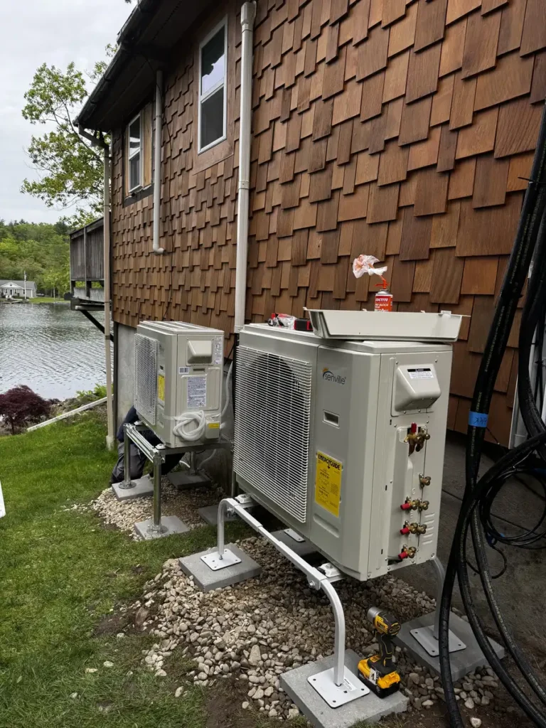 A white multi-zone mini-split air conditioner condenser cleanly installed on ground brackets outside a home with brown cedar shake siding in the Capital District, NY.