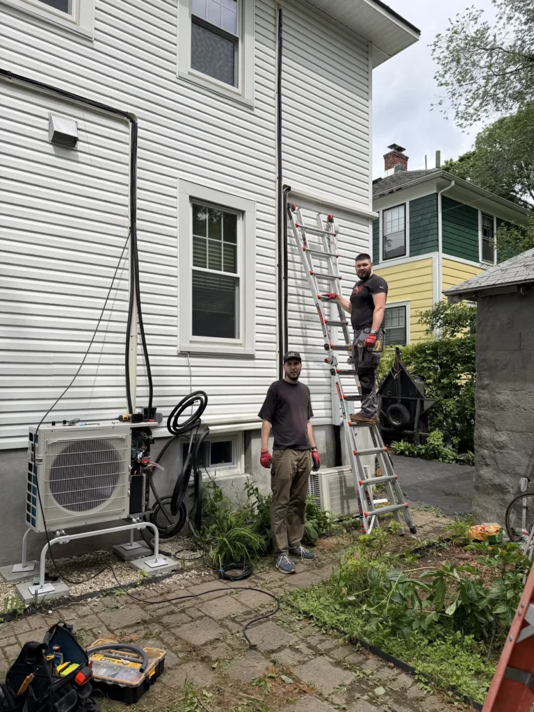 Two Astello LLC technicians stand outside a white two-story residential house during an HVAC installation in Albany, NY