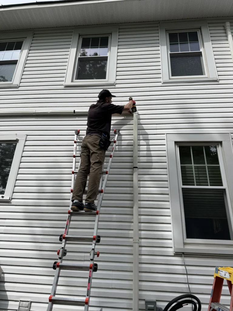 An Astello LLC technician wearing a black Caterpillar baseball cap and red work gloves stands on a ladder, using a cordless impact driver to secure a white vertical line hide cap onto the beige vinyl siding of a house. Troy,NY