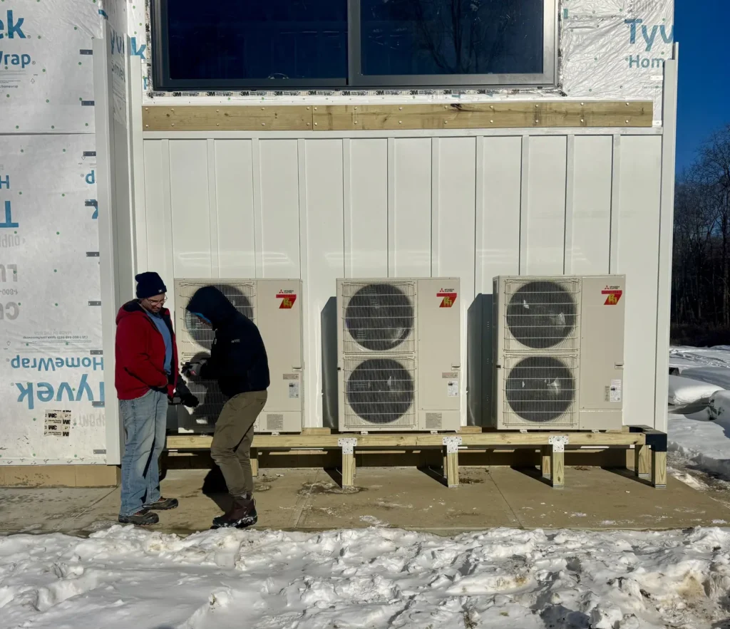Astello LLC technician checking the wiring on a bank of four Mitsubishi Electric hyper-heating mini-split condensers installed outside a building on a wood mounting stand during winter in Albany, NY.